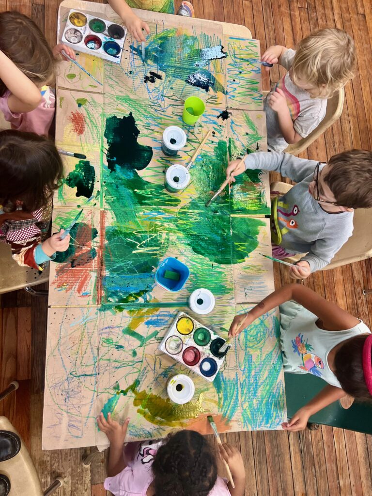 A table full of pre-K children sitting in kid-sized chairs to paint together on a large flat surface. Each kid has their own brush and access to many different paint colors; the painting is predominantly green.