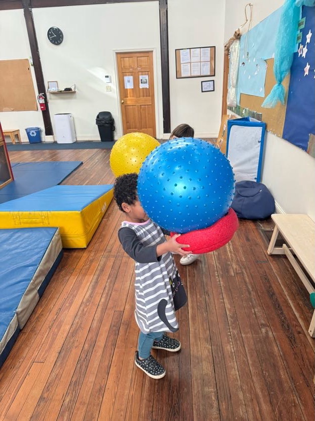 A child carrying two large balls in the indoor gross-motor play space--their face is hidden behind their armload! Colorful play mats are visible beyond.