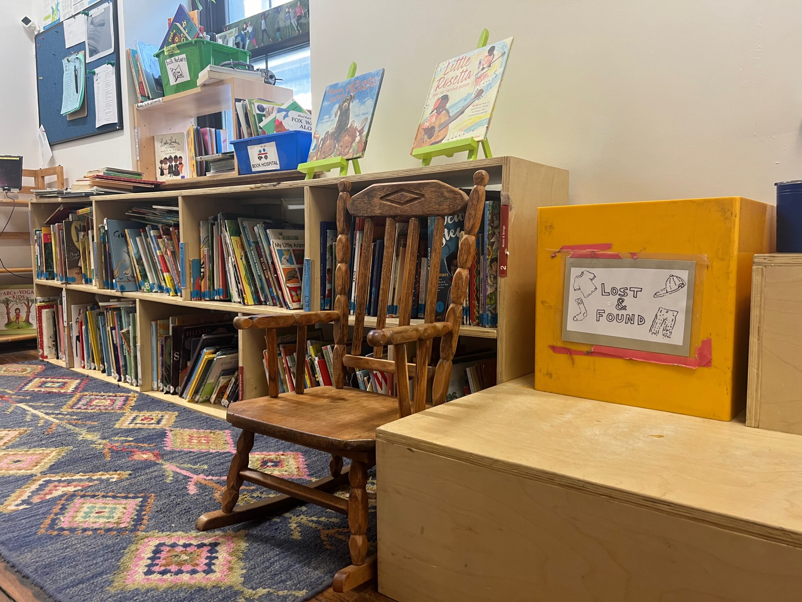 A wooden child-sized rocking chair tucked against full bookshelves in the library. Some books are on display above the shelves.