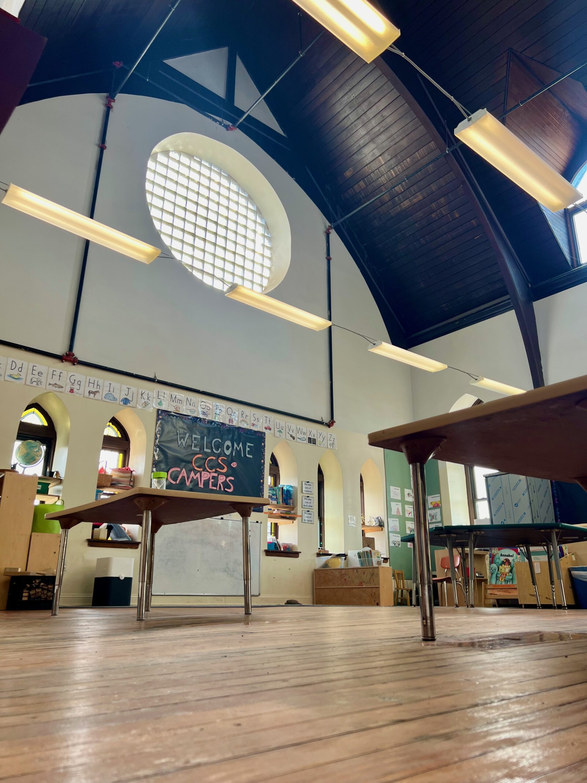 A pre-K classroom with high, arched ceilings and tall, arched windows. Child-sized tables and chairs and colorful wall decorations and books are visible.