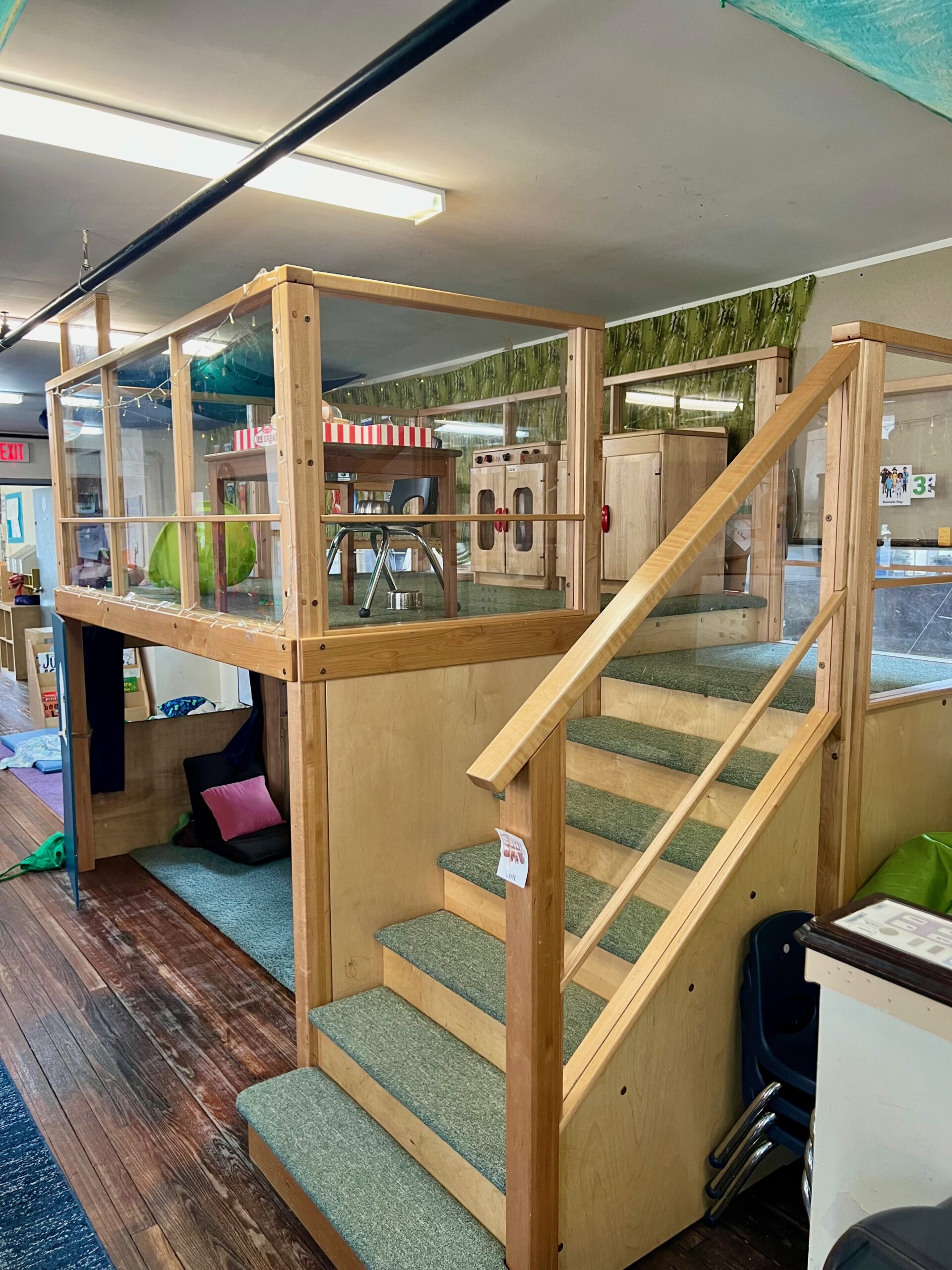 A lofted playspace in a pre-K classroom, with carpeted stairs and a protective railing leading to a small toy kitchen and cafe area for imaginative play. A cozy nook with rugs and pillows is visible underneath.