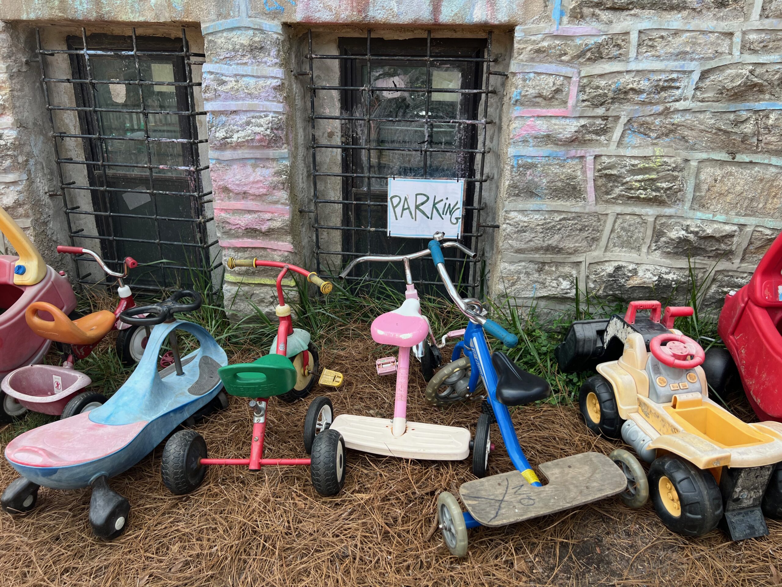 A range of toddler-sized tricycles and toy vehicles gathered in the outdoor play-yard by a handwritten sign reading "Parking."