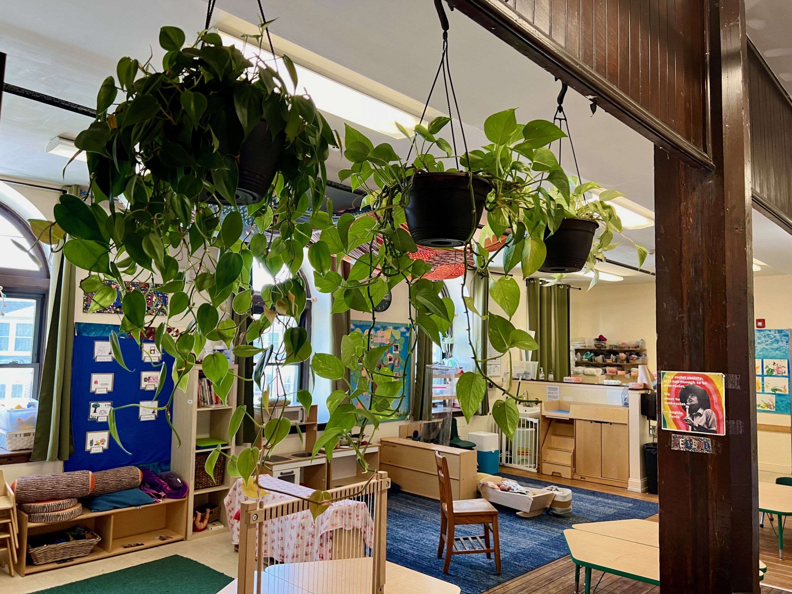 Trailing green plants hanging high up in a pre-K classroom, with a play kitchen, child-sized chairs and tables, and toys and decorations visible beyond.