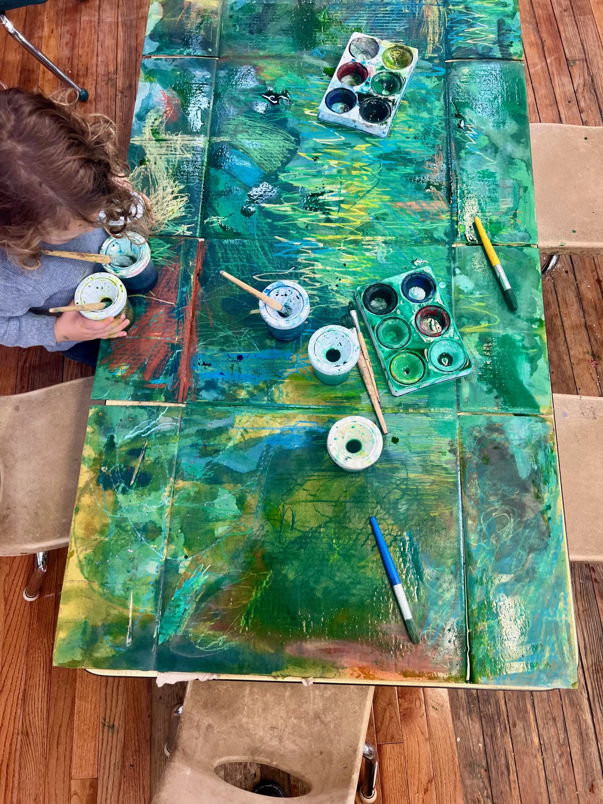 A child leans over an art project painted in many shades of green, with accents of yellow, blue, and brown. They are holding two paint containers with brushes and many other paint palettes and brushes are scattered on the surface. Child-sized chairs are gathered around.