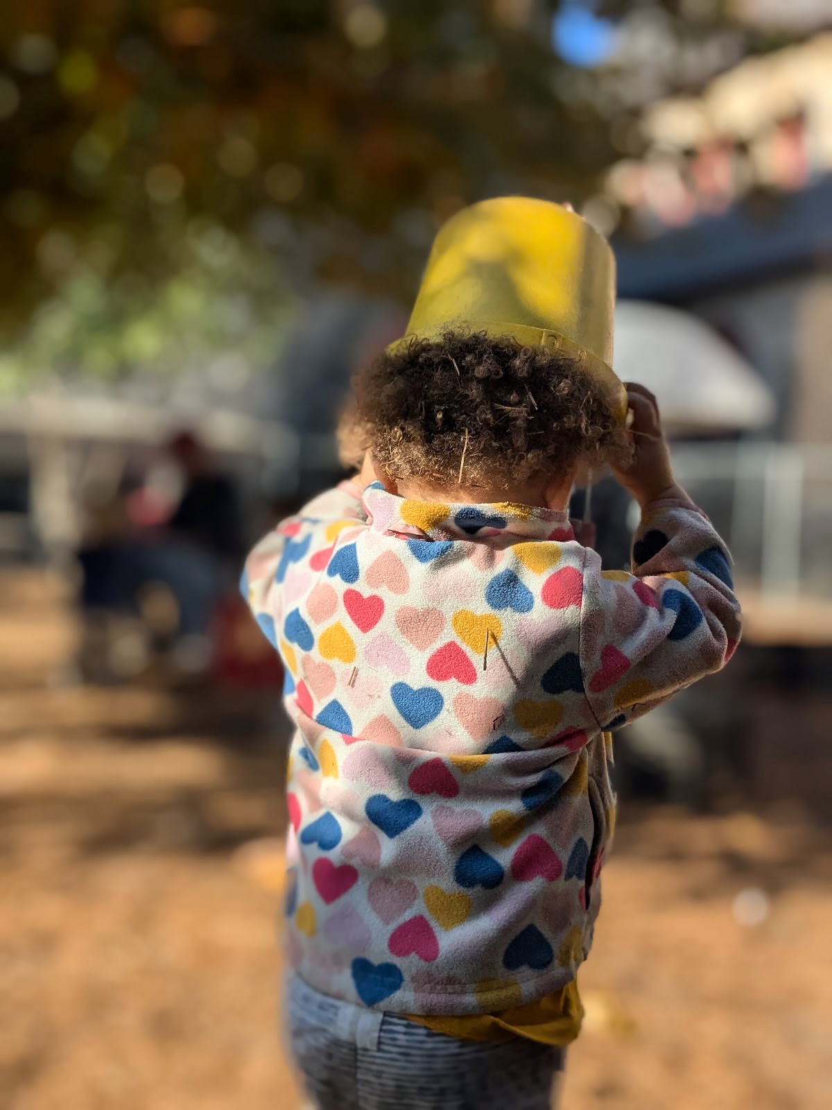 A toddler in the outdoor play-yard fitting a yellow bucket over their head like a hat.