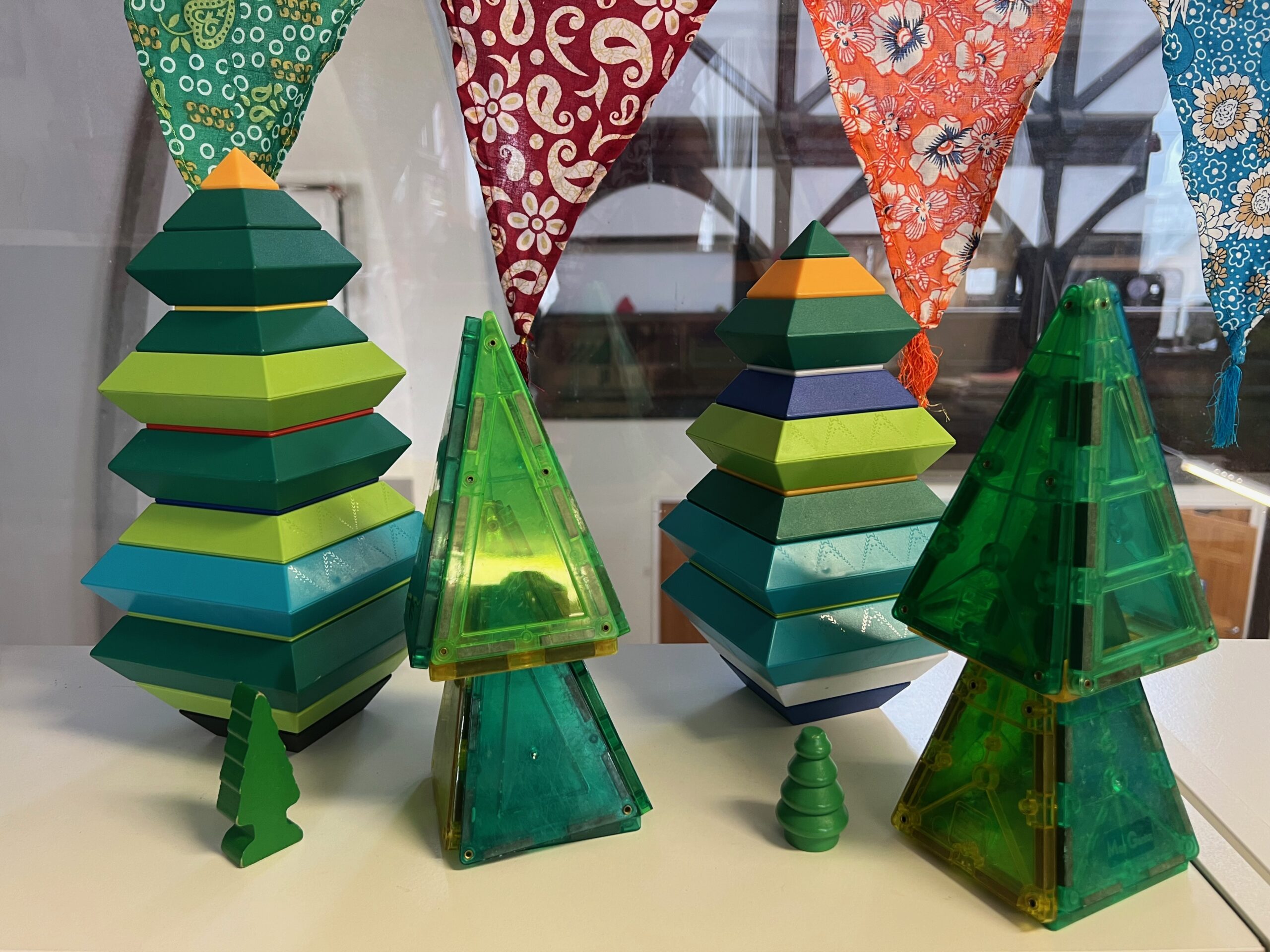 A display of tree-inspired constructions built of magnatiles and other blocks, mostly green in color. The "trees" are sitting on a white desk in front of a window, with a colorful patterned pennant hanging behind.