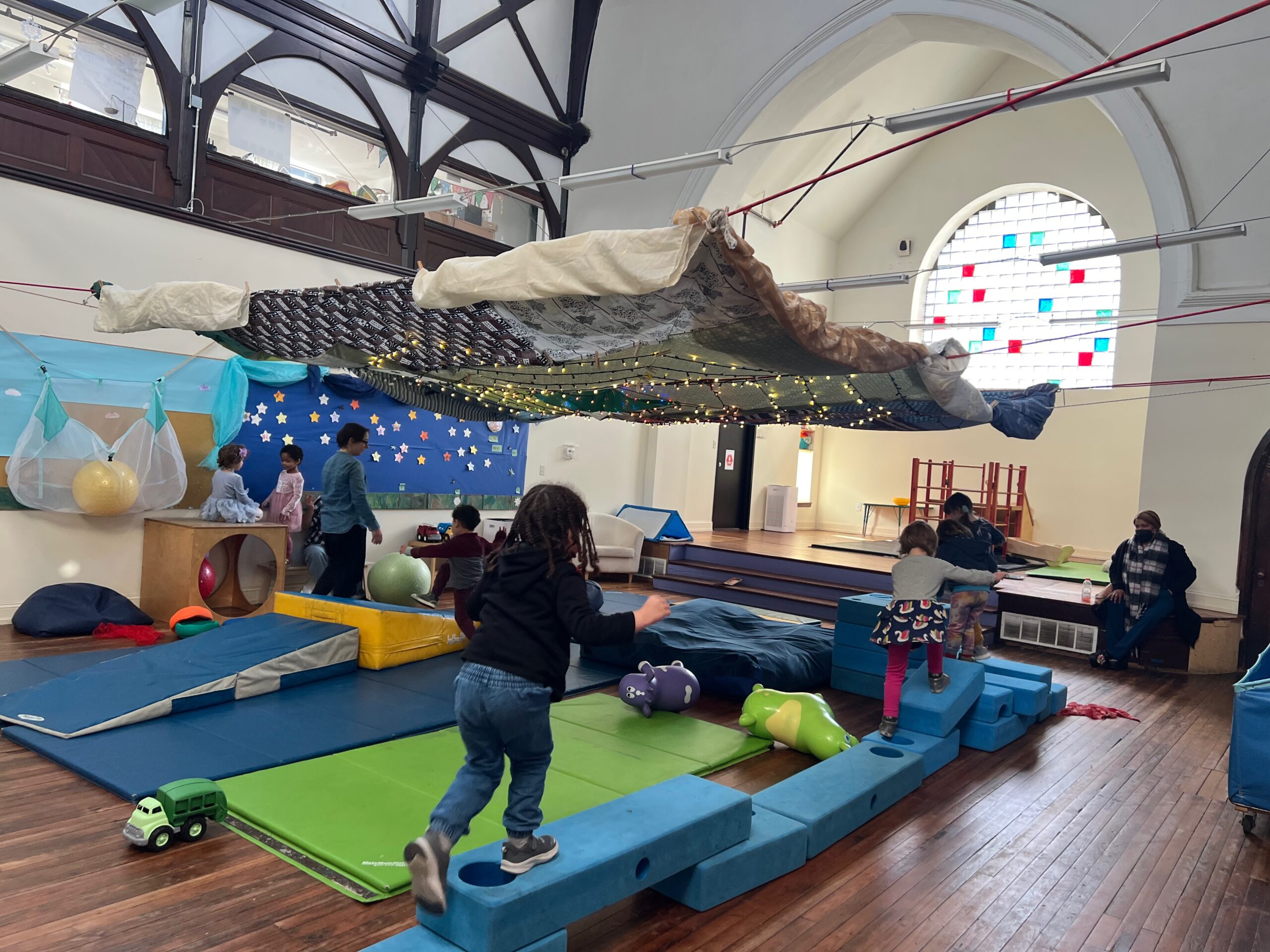 Pre-k children and teachers playing in the indoor gross motor play space. Some kids are using large blue foam blocks as a giant balance beam, while others are climbing on ramp-shaped pads, giant exercise balls, and a wooden climbing structure. Colorful playmats are gathered together on the wooden floor. There is lots of light coming through the arched stained-glass window at the back of the room, and all the activity is happening below a festive overhang of blankets and twinkle lights.