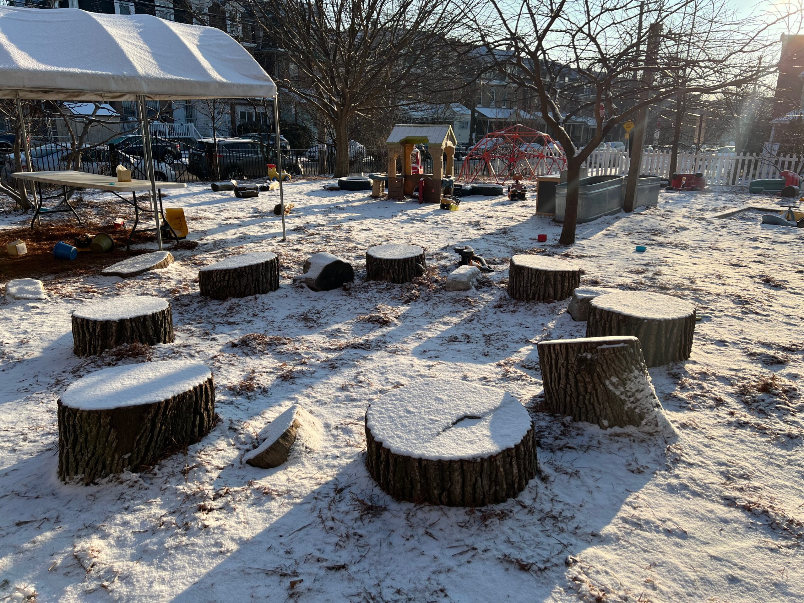 The outdoor play-yard lightly covered in snow on a sunny winter morning. A circle of snow-covered tree stump pieces is in the foreground, while a shade tent, play house, and climbing structure are visible beyond, interspersed with small trees and large planters.